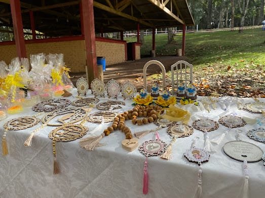 Mesa comprida coberta de artesanato do ateliê, montada no gramado, sob o galpão