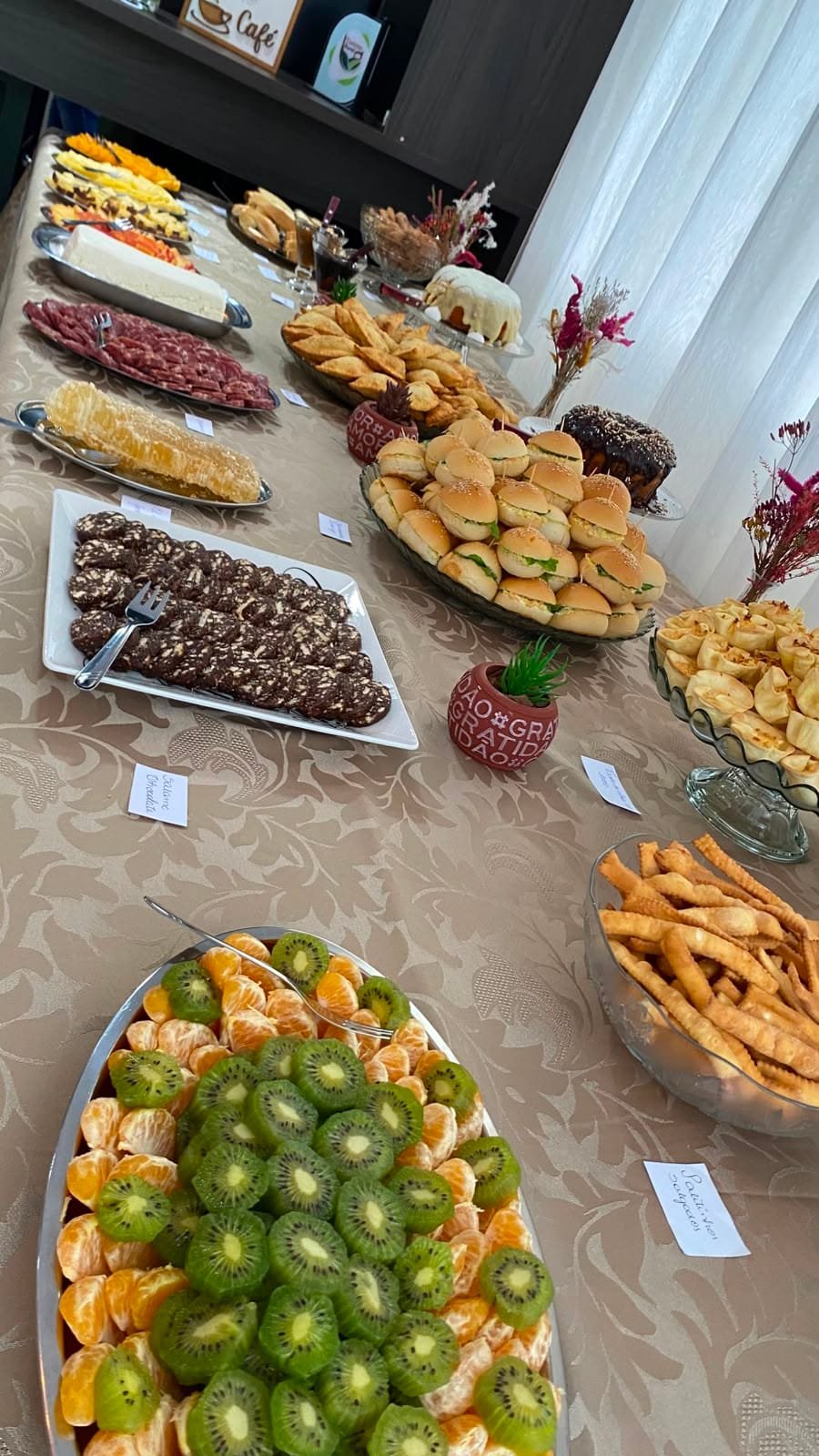 Mesa do café colonial com salgados, doces, frios e uma travessa de kiwi e laranja