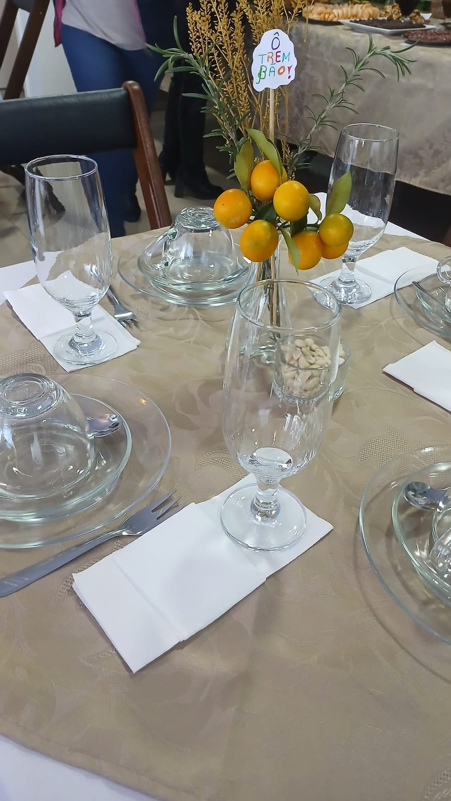 Mesa posta no salão, com taças, pratos de vidro e um arranjo de laranjinhas