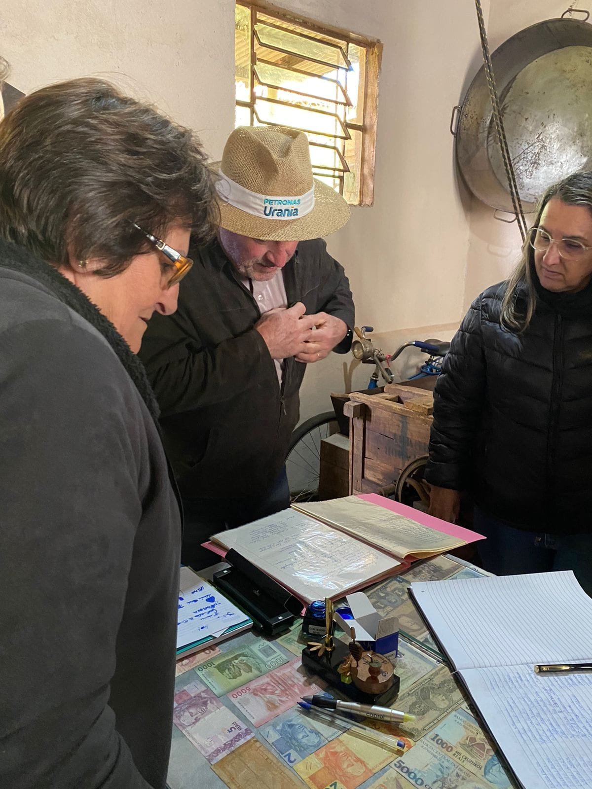 Visitantes debruçadas sobre documentos e cadernos antigos, com o anfitrião de chapéu ao lado