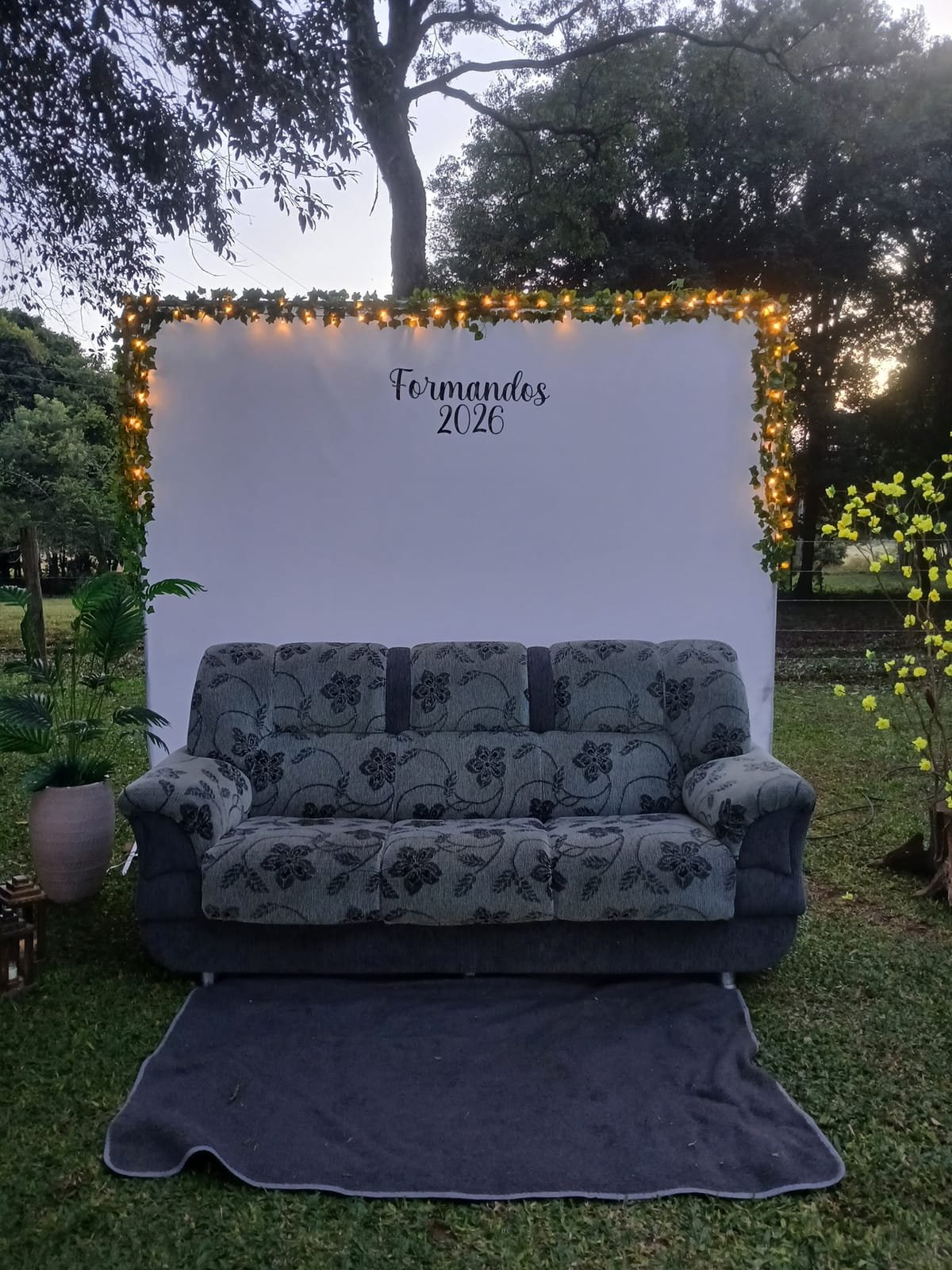 Painel branco escrito Formandos 2026, com luzinhas e um sofá na frente, montado no jardim